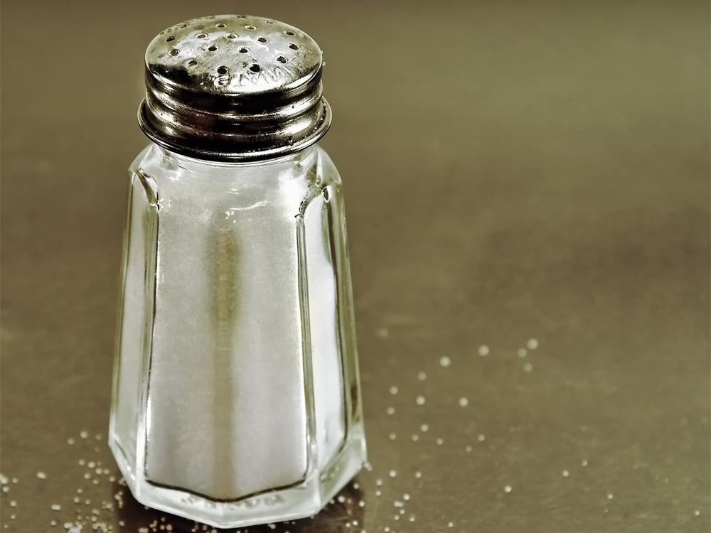 A close-up of a full salt shaker sitting on a counter. A few grains of salt are sprinkled around it.