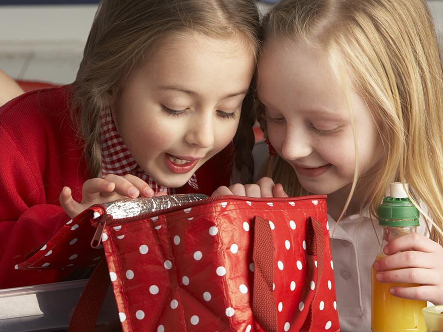 Two girls peer into a lunchbag. One of the girls holds a drink in her left hand.
