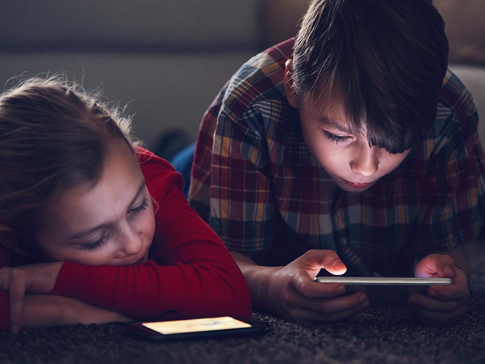 A young boy and girl lay on the floor, each looking at a cell phone screen. The boy holds his phone, the girl’s phone is laying on the floor in front of her, and she’s resting her chin on her arms.
