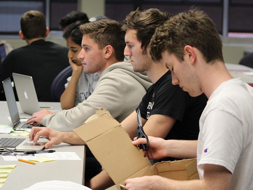 Three people sit at a table and look at computers while another person cuts a cardboard box. 