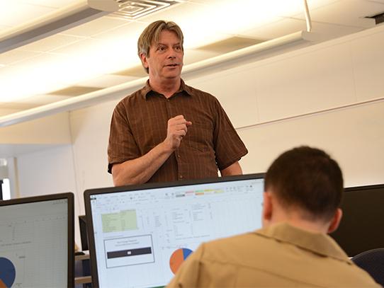 A standing person talks to a person sitting down at a computer. 