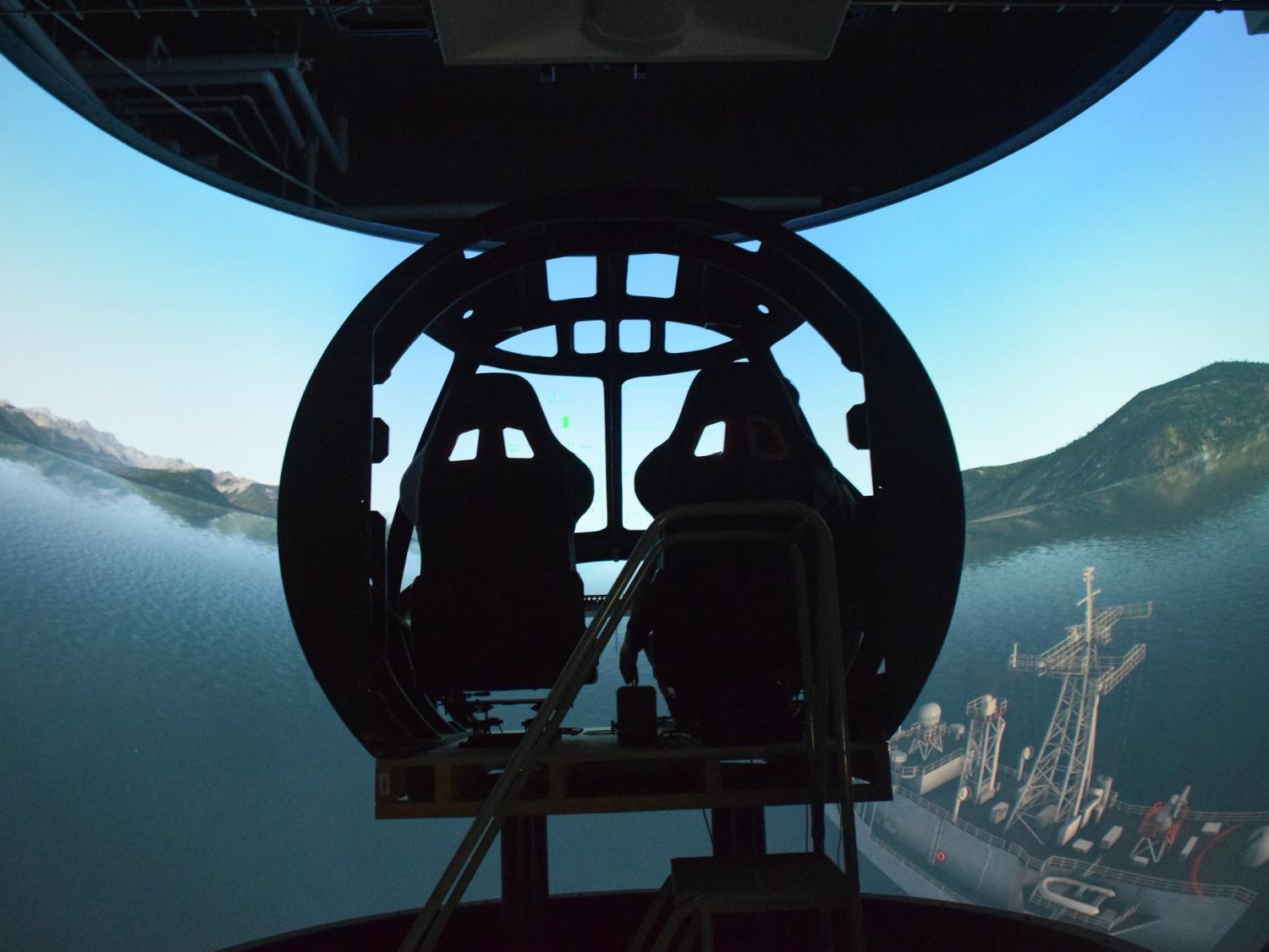 A cockpit is shadowed against a simulated water scene over a ship with mountains in the background. 