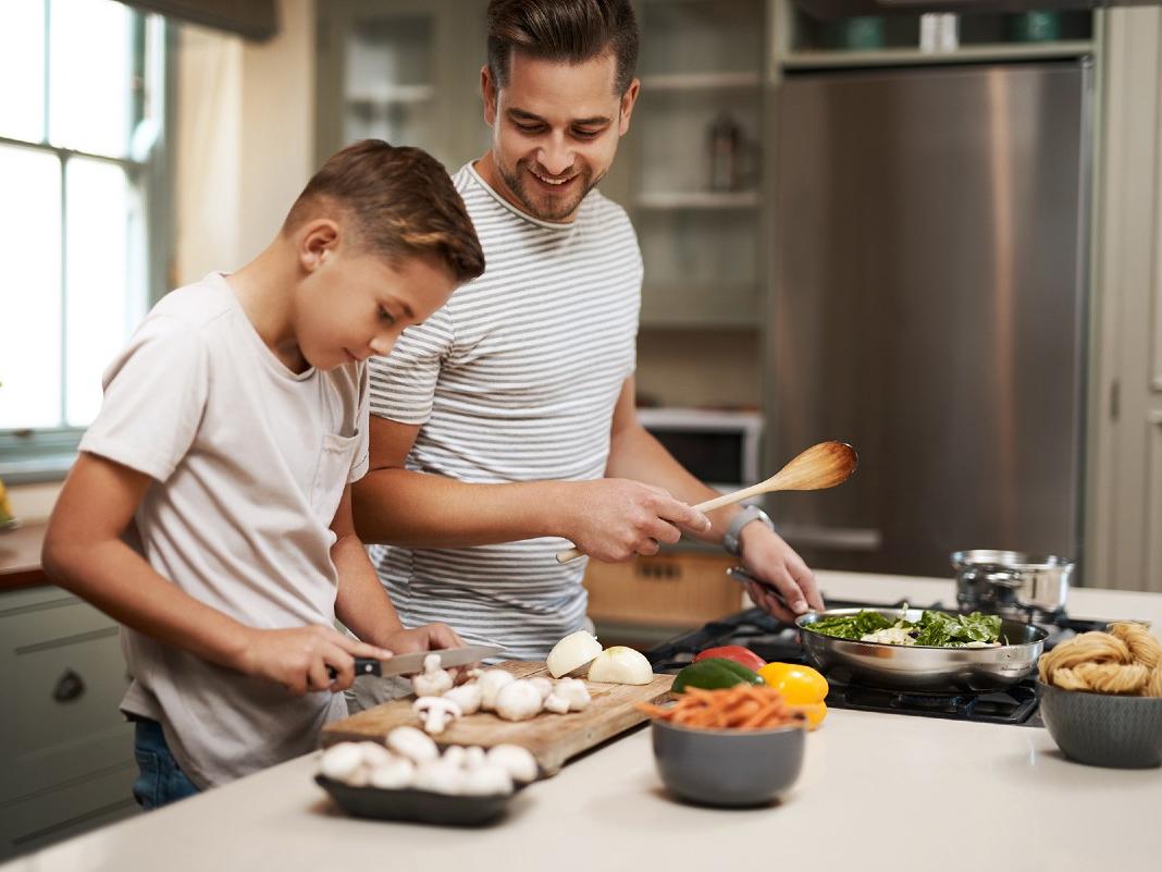 A father prepares dinner while watching his son chop mushrooms. 