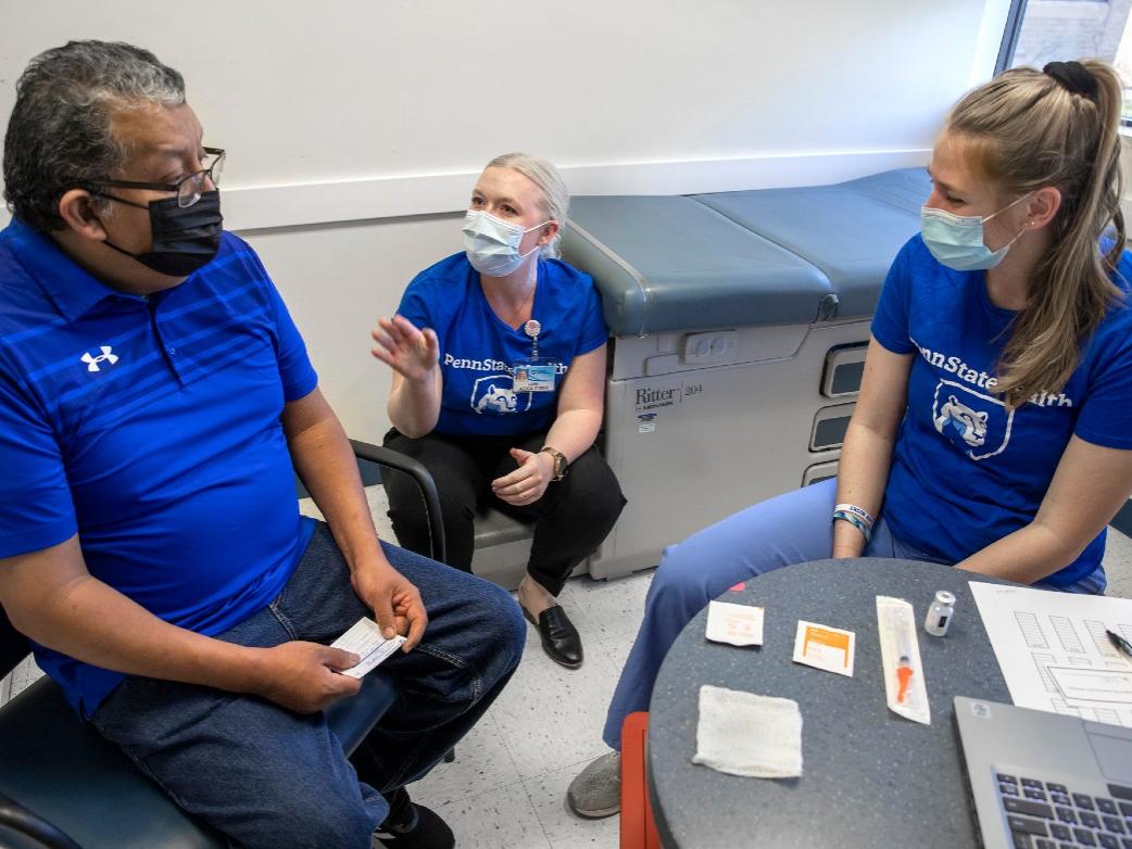 Three people - a man and two women – sit in a clinical room, talking. A laptop computer is on a table in the foreground.