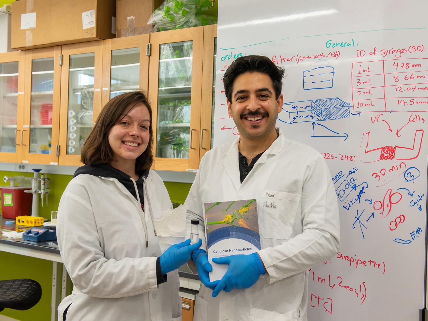 A man and woman in lab coats smile in front of a white board. The woman is holding paper, while the man holds a book.