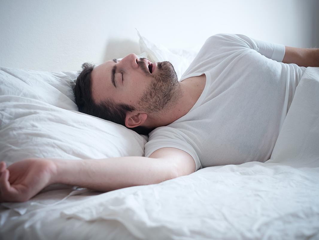 A sleeping man snores while laying on a bed. 