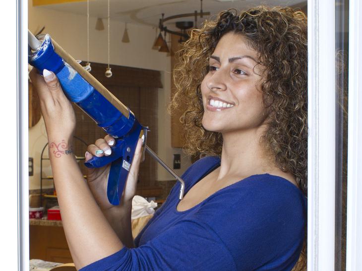 A woman stands with a caulking gun by a window