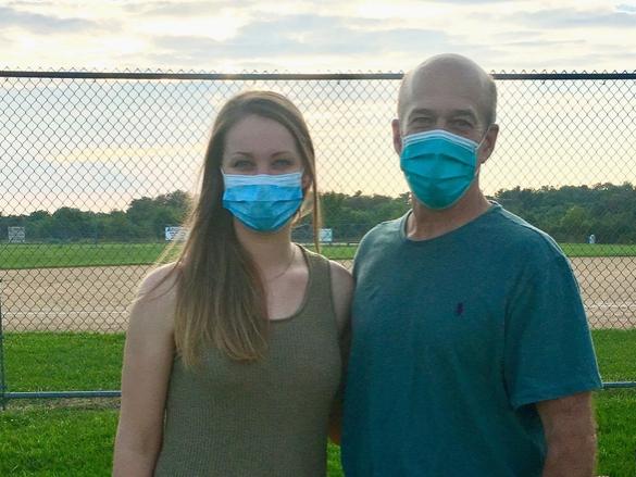 Shalisa Walker stands arm in arm with John Dohner in front of a chain link fence at a softball field. Walker has long hair and wears a tank top. Dohner, who is bald, wears a T-shirt. Both are wearing masks.