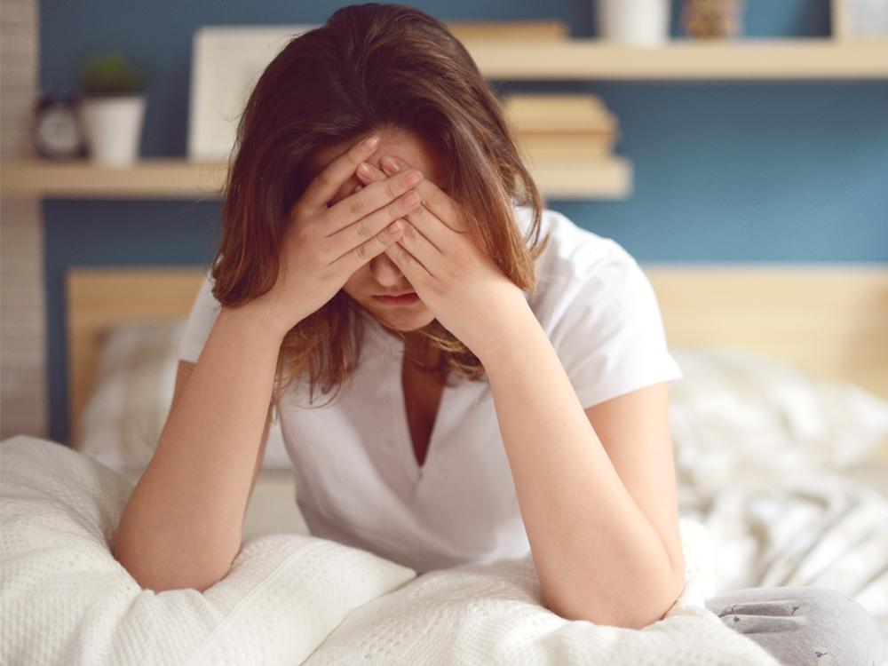 A woman sits up in bed, leaning slightly forward, with her head placed in her hands.