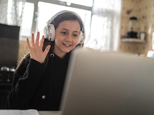 A girl wearing a sweatshirt and large headphones smiles and raises her hand in front of a laptop.