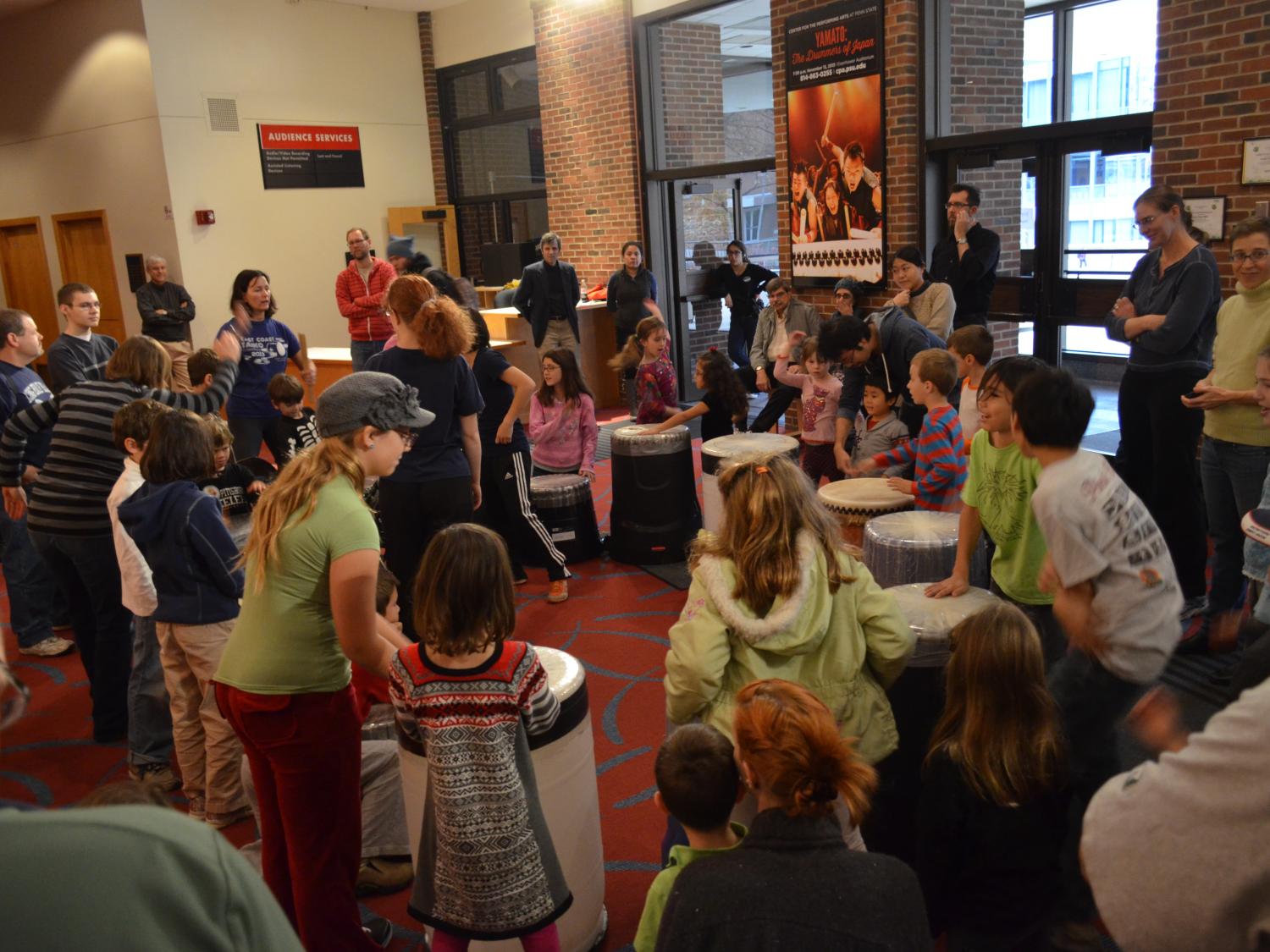 People of all ages crowd around various large drums while they listen to a workshop leader.