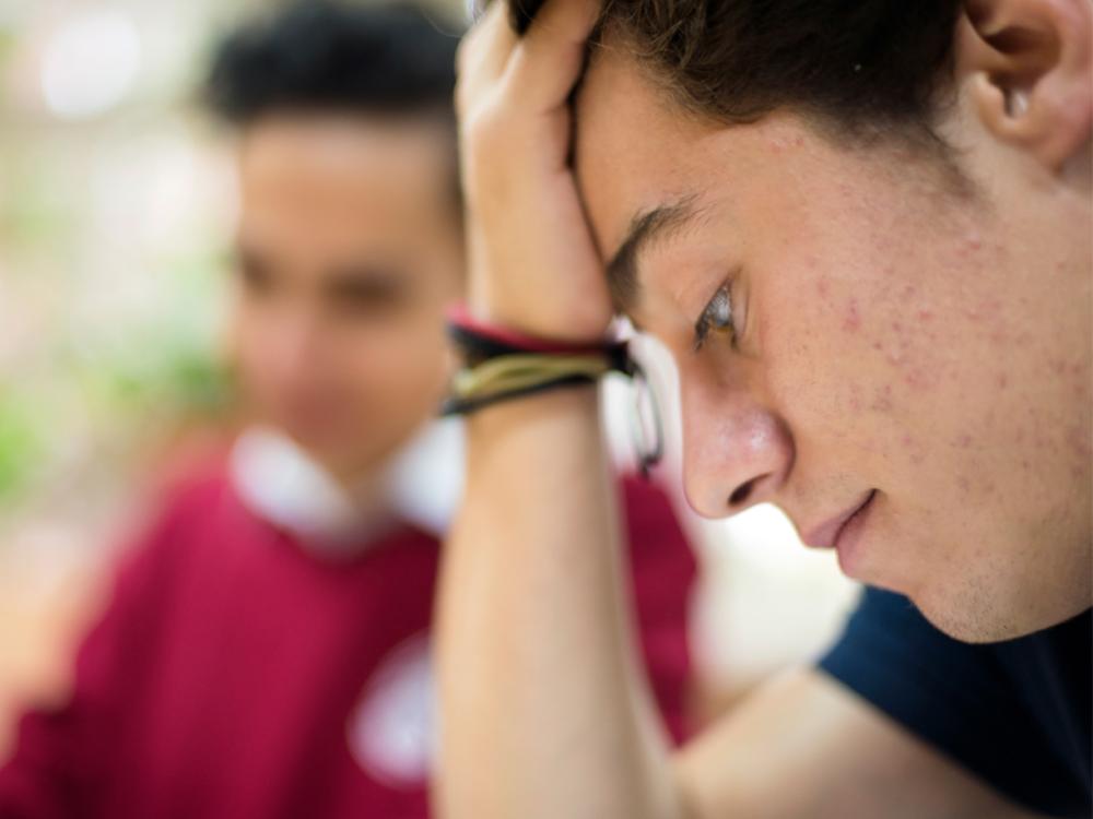 A close-up side view of a teenage boy with his head resting on his hand, looking down. Acne is visible on the side of his face.