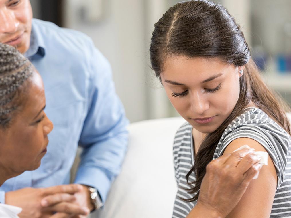 A medical professional in a white coat wipes the left arm of a teenage girl, apparently in preparation for a vaccination. The girl looks down at her arm. A man is seated nearby, looking on.