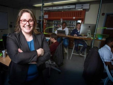 Woman in suit jacket stands in an empty lab as she smiles before camera.