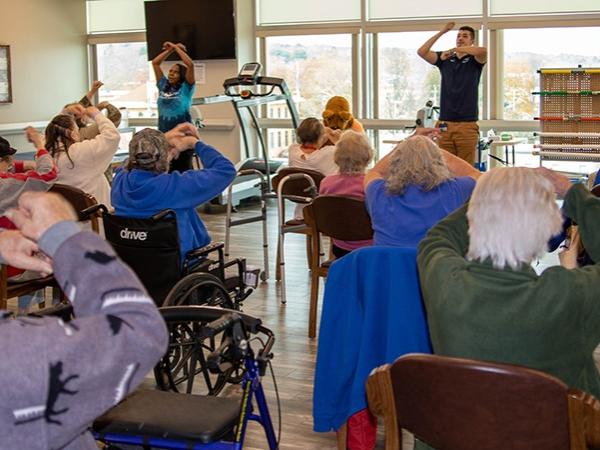 A group of seniors complete exercises in their chairs while instructors stand in front of room