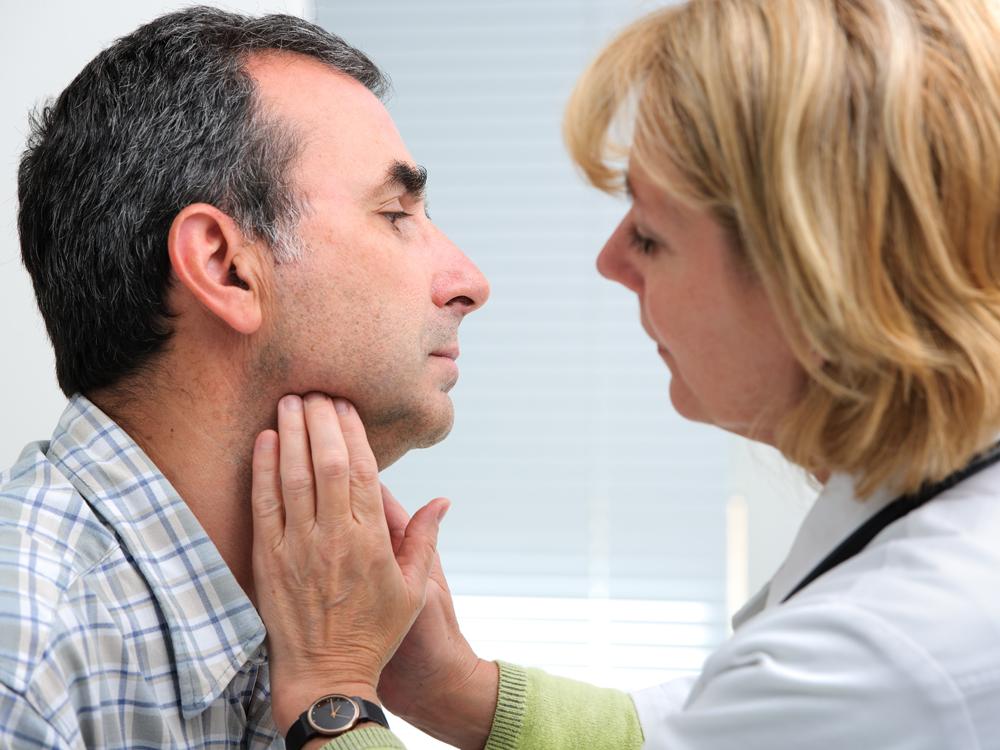 A female physician wearing a white coat and stethoscope places both hands on the throat region of a male patient.