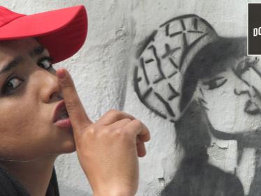 teenage woman wearing a red baseball cap with her index finder covering her mouth to show the sign for quiet