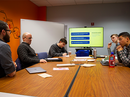 Five men sit around a table and look at a large screen. 