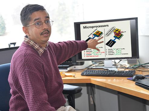 A man wearing glasses and a maroon sweater gestures toward a computer screen displaying information about microprocessors while he talks to someone off to the side of the camera. 