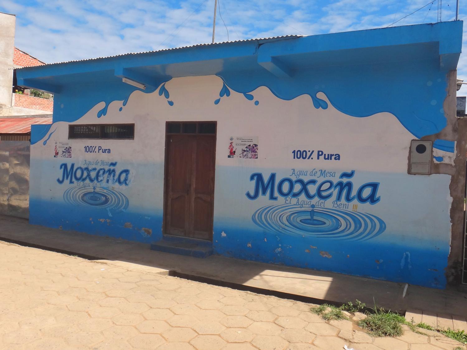 A small shop that sells water on a dirt road situated among other buildings along a dusty, unpaved road