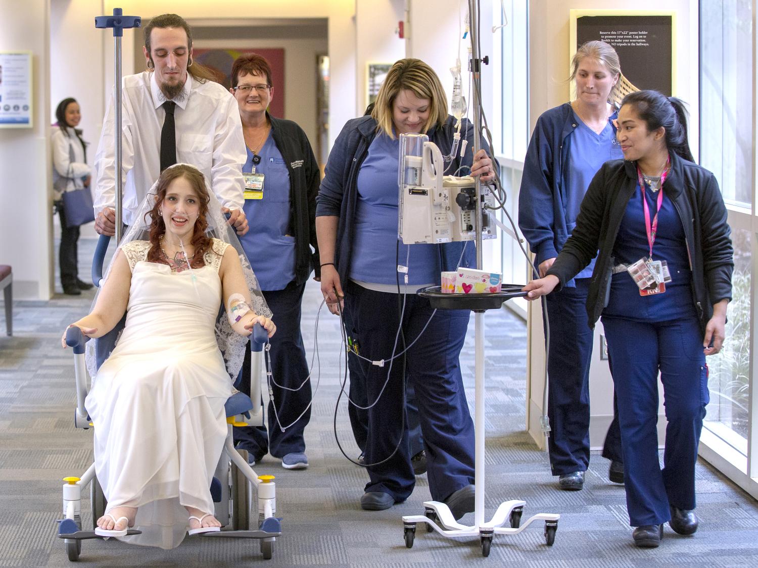 A man in a white shirt and black tie pushes a woman in a wedding gown in a wheelchair down a hospital corridor. With him are several members of the hospital staff in blue nurse’s uniforms. 
