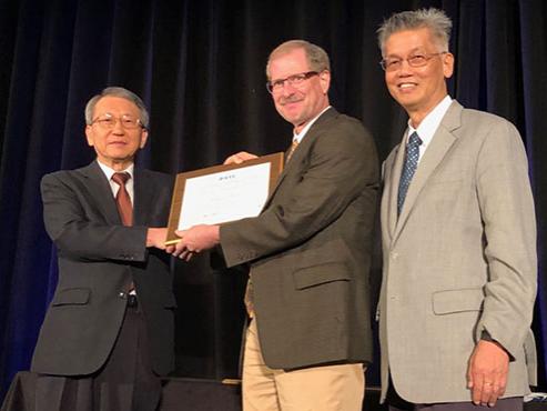 Two men shake hands as one man hands the other man a plaque. A third man stands by. 