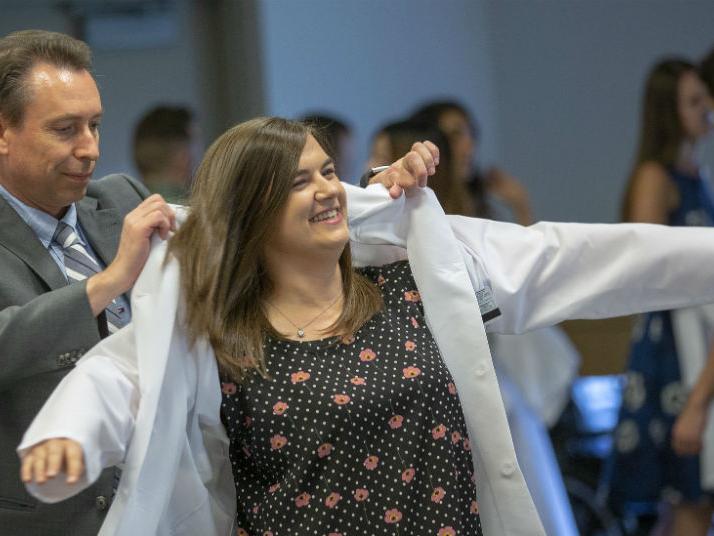 Ralph Keil, left, director of the biomedical sciences graduate program at Penn State College of Medicine, helped a woman graduate student put on a lab coat during the Graduate Student Oath Ceremony. Behind them are other students.
