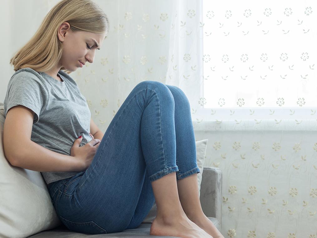 A woman dressed in jeans and a t-shirt sits on a couch. She looks down, and both of her hands are placed on her abdomen.