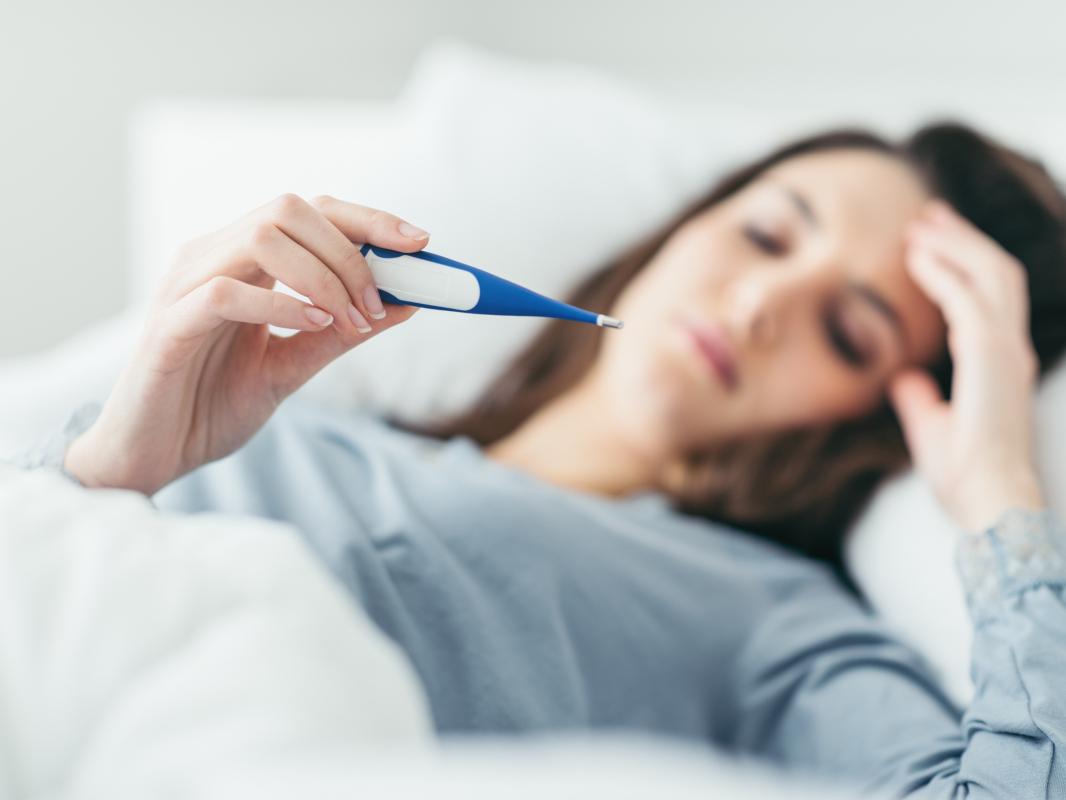 A woman lays in a bed, resting her head on her left arm. Her face is slightly out of focus. She holds up a digital thermometer and looks at it.
