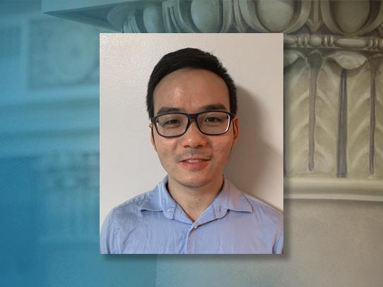 A photo of a smiling man with dark hair wearing a blue shirt and glasses on a background featuring a close-up of a classical column capital
