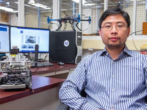 A man wearing glasses and a blue, vertical-striped shirt poses in front of his desk. On the desk to his right is a small device with wheels and wires, and behind him on the desk are two computer monitors and another small robot-like device. 