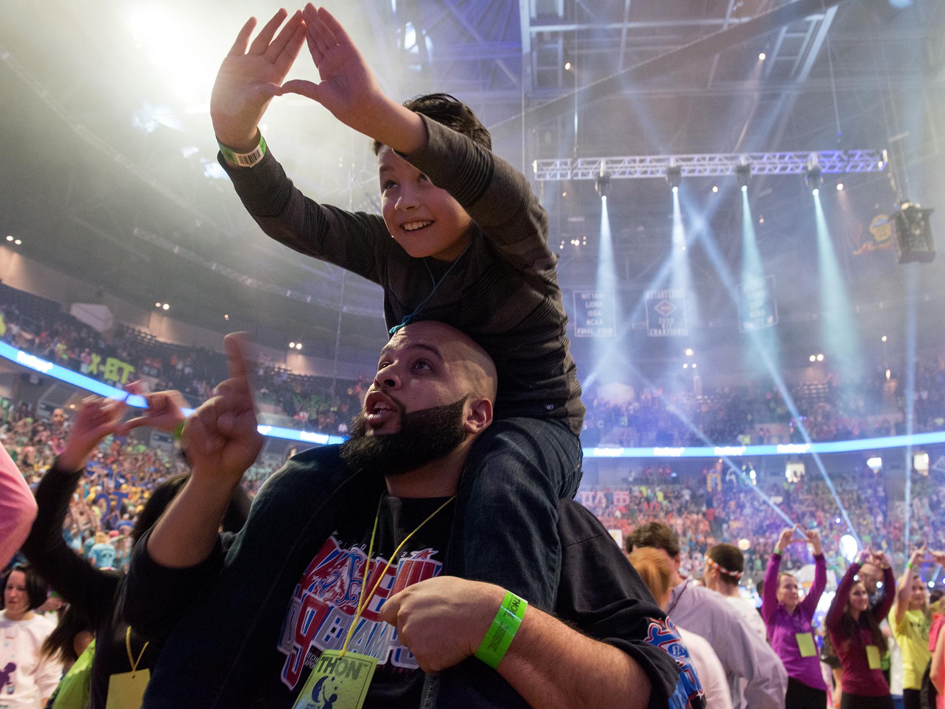 Boy on his father's shoulders making a diamond sign at THON 2015