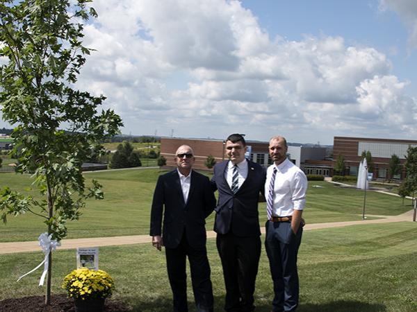 Three men stand on campus beside a commemorative maple tree to honor the memory of Trevor Thomas.