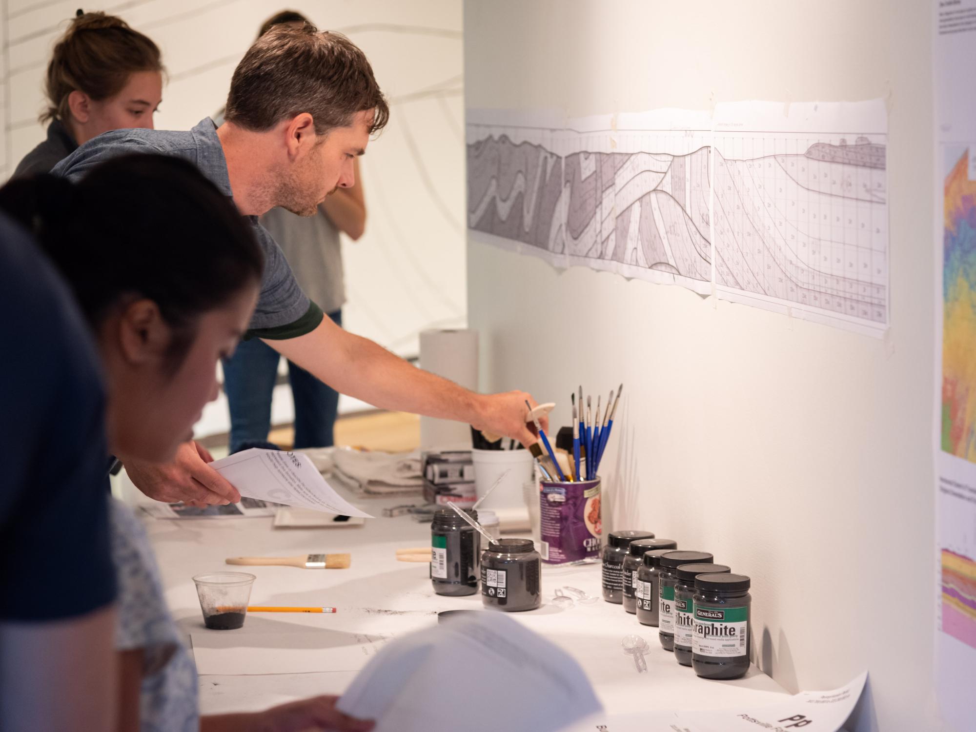 Spangler reaches for a paint brush on a table with drawing materials among volunteers