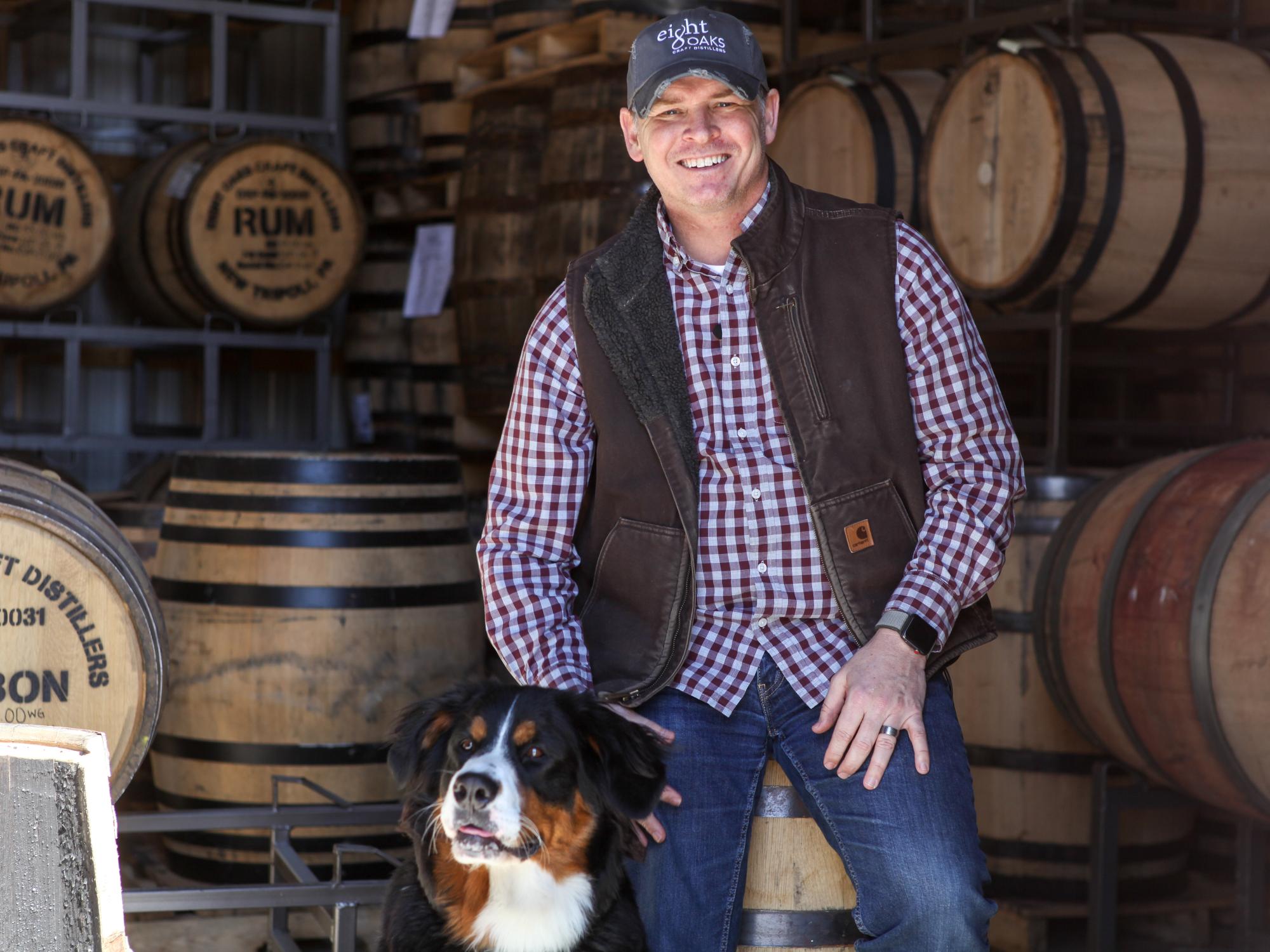 photo of man in vest with dog surrounded by barrels