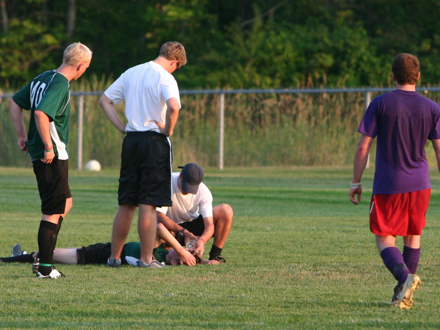 Injured soccer player laying on his back on the pitch. Concerned coaches attend to him as two players look on, concerned