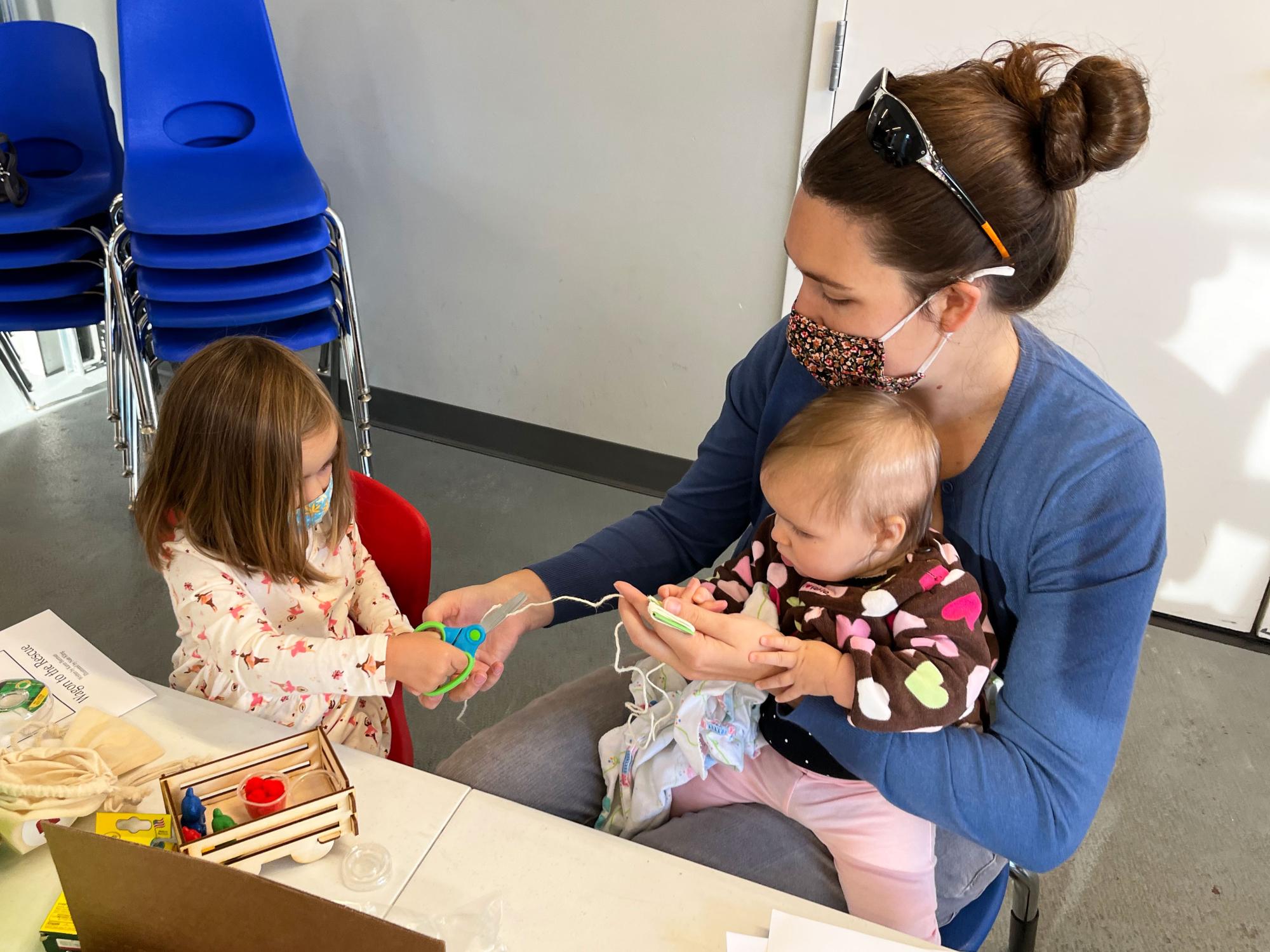 Mom and young daughter cut a string while baby on mom's lap looks on