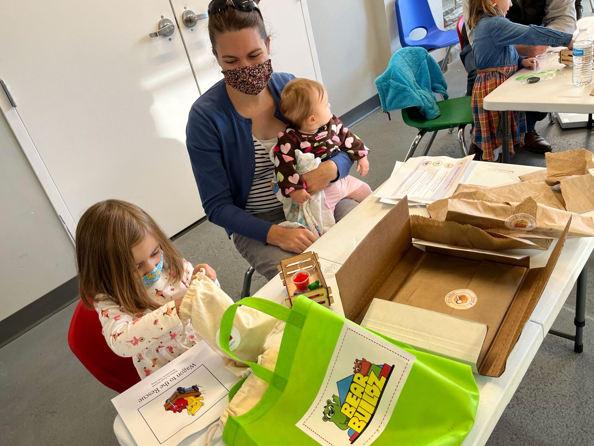 Young girl opens a bag of Bear Buildz building materials next to her mother and baby sister