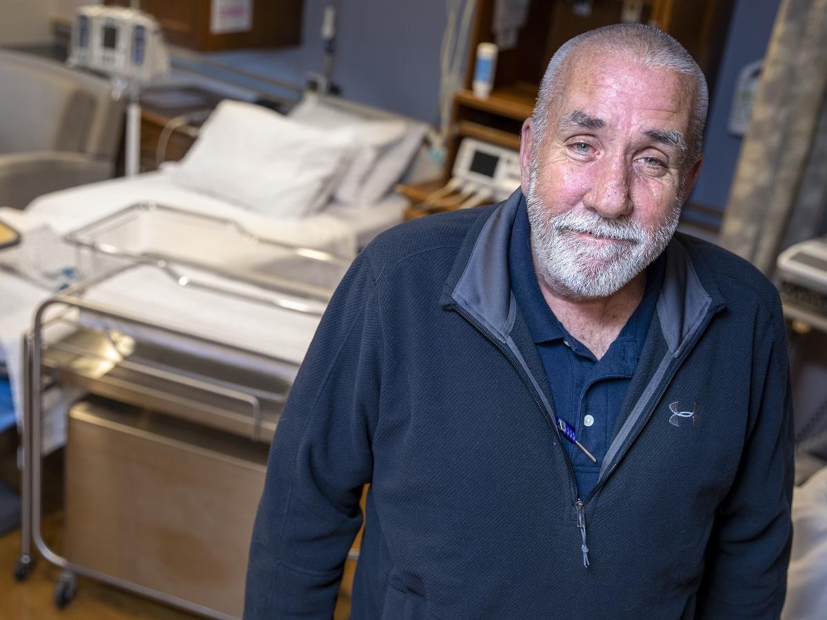 Matt Taylor, who has a crewcut, beard and moustache and wears a quarter-zip jacket, smiles as he stands in front a bed and bassinet in the labor and delivery unit at Holy Spirit Medical Center.