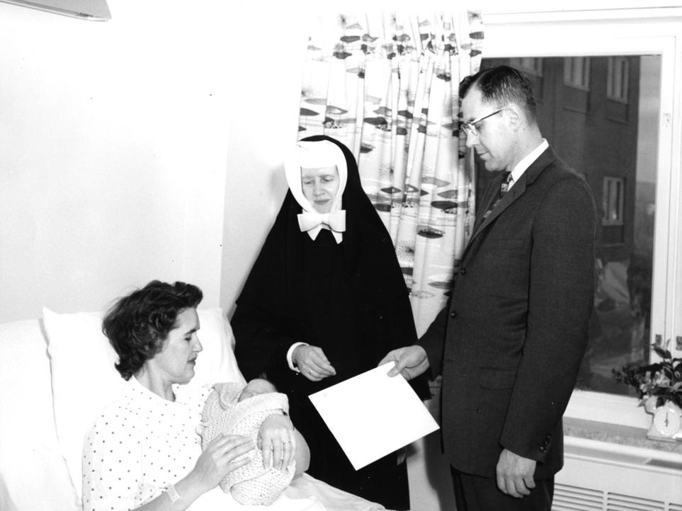 Esther Taylor, who has short hair and wears a hospital gown, lies in a hospital bed, cradling her newborn son, whose face is peeking out from a blanket. A nun and a man in a suit holding a large envelope stand by the bed.