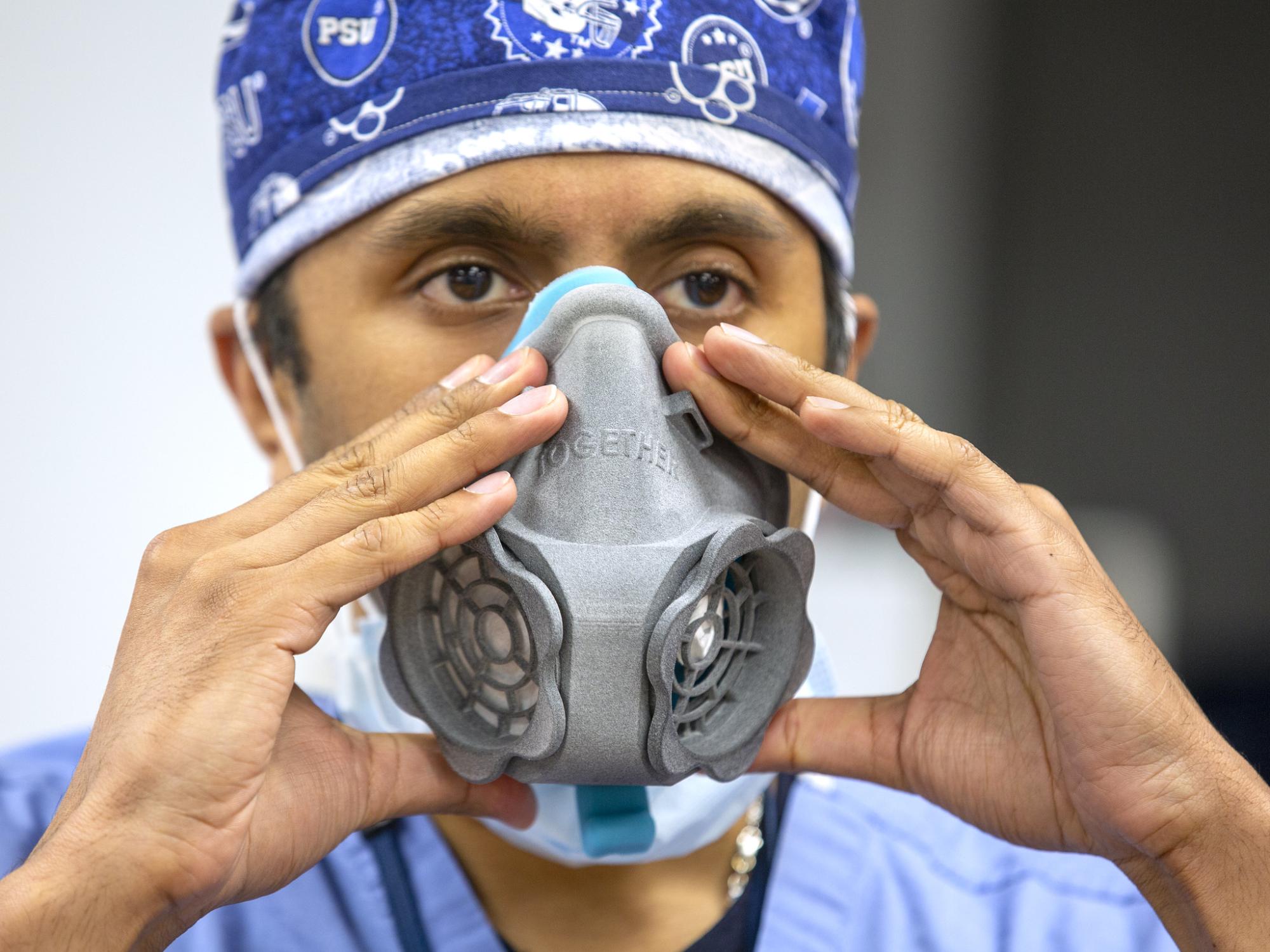A man holds a 3D printed N95 respirator up to his face with both hands. He wears surgical scrubs and a head covering bearing Penn State athletic symbols.