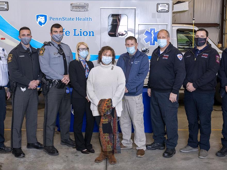 Kate Warnigiris, wearing a bulky sweater, jeans and a mask, is surrounded by eight people who helped save her life during a cardiac arrest. Crew members, who are wearing uniforms from their respective agencies and masks, stand in front of a Life Lion ambulance in a garage.