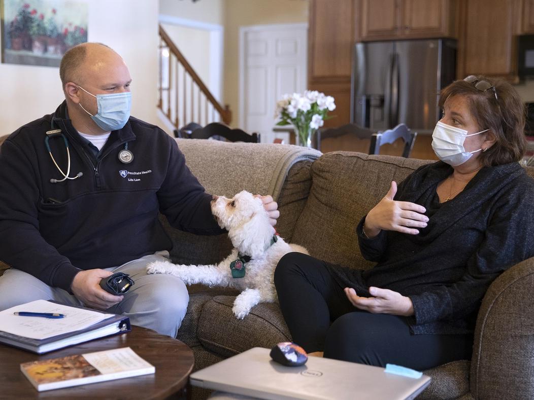 Kate Warnigiris, who has short hair with glasses on the top of her head and wears a shirt, pants and face mask, gestures as she talks with Loren Miller on a couch in her living room. Miller, dressed in a Penn State Health jacket with a stethoscope around his neck and a face mask, pets a dog on the couch as he listens.