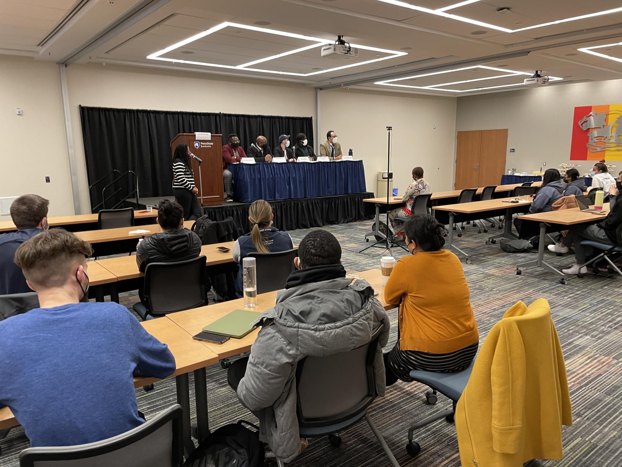 Community members seated at long wooden tables watch a group of panelists sitting on a stage with a facilitator standing in the foreground. 