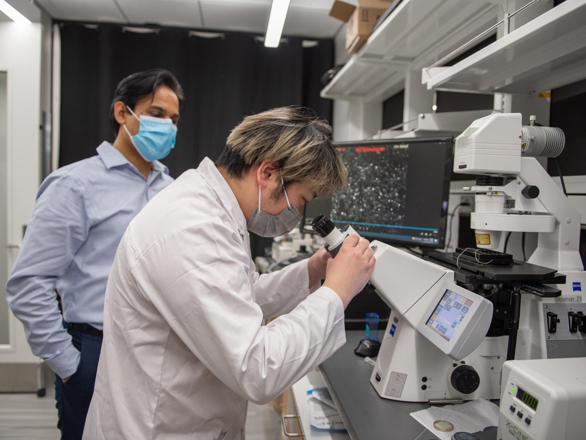 Two men stand side-by-side, as one stares into a microscope. 