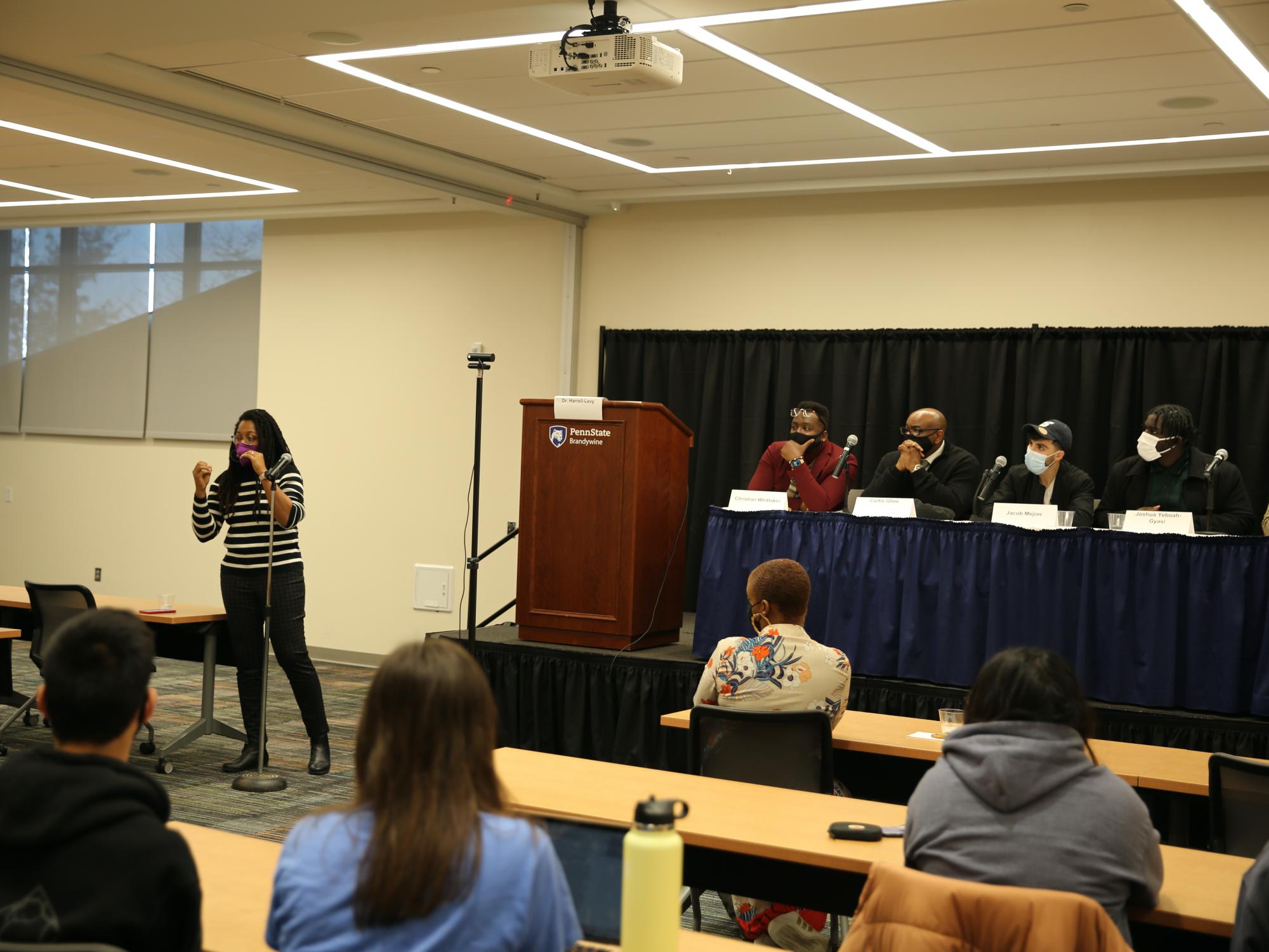 Panelists sit on the stage at a long table while a moderator facilitates a conversation with students, faculty and staff.  