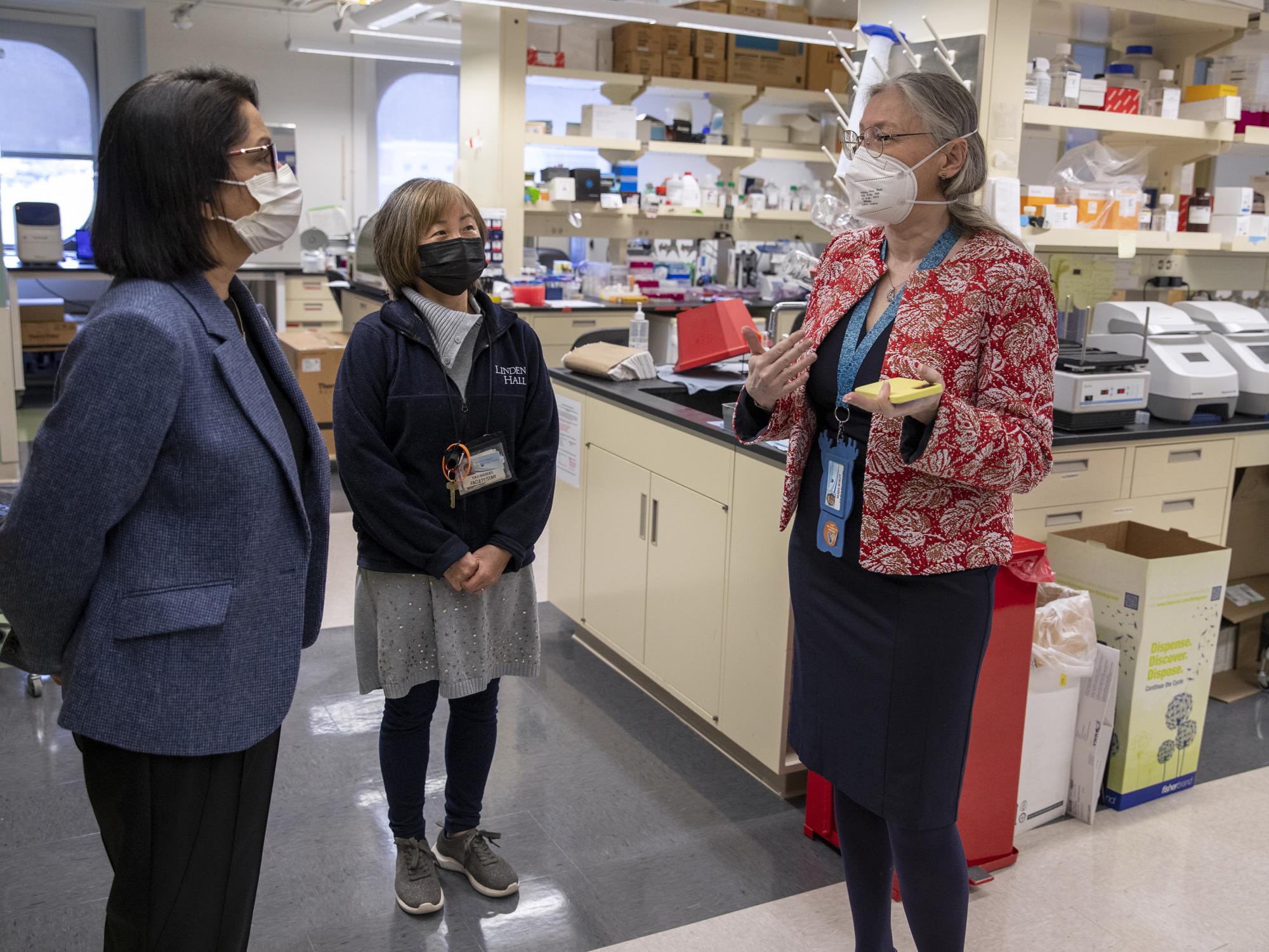 Three women stand in a lab, talking. All three wear face masks. Lab equipment is in the background.