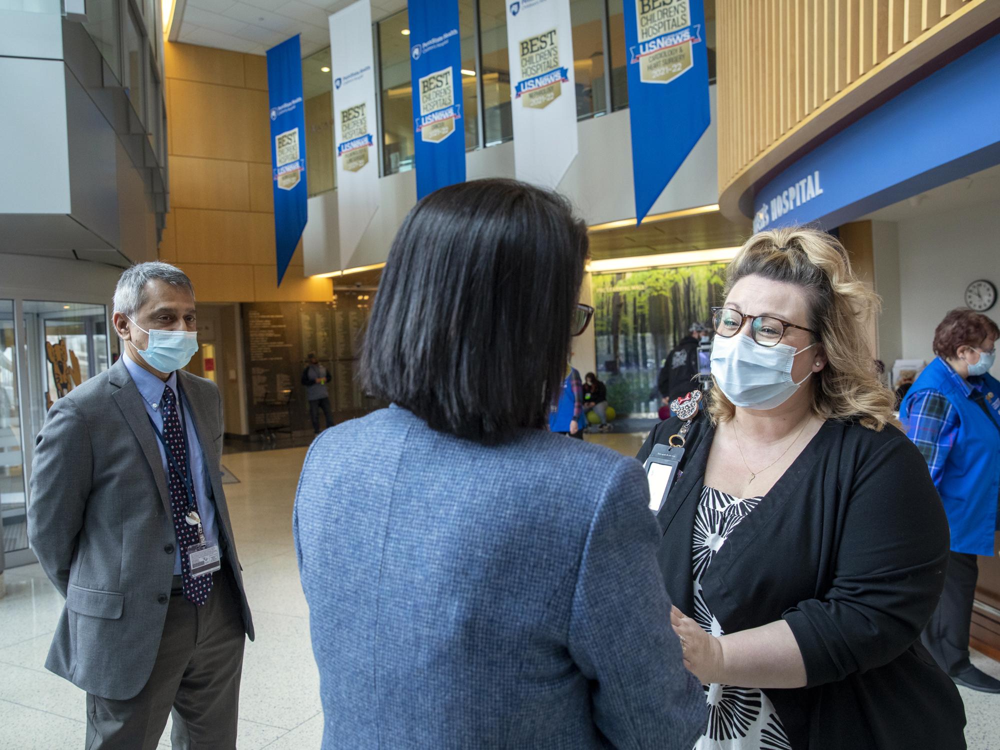 Two women talk and shake hands. A man stands at left, looking on. They are in the lobby of a building.