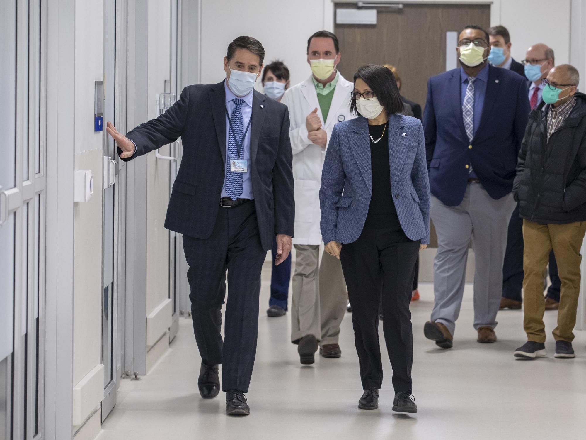A man and woman in professional attire walk down a hospital hallway, as the man points to a room on his right. Several people are walking close behind.