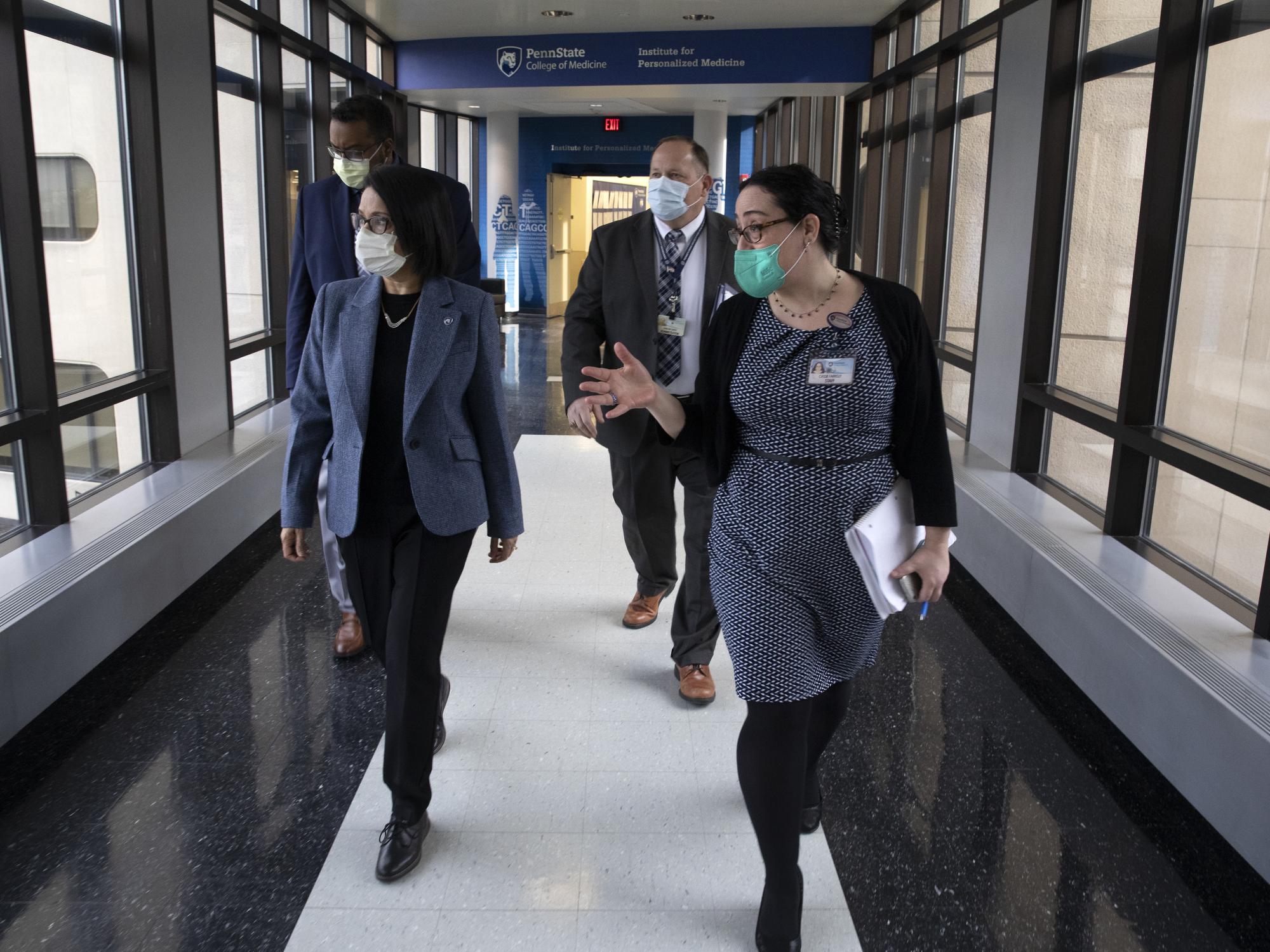 Four people walk down a hallway with windows on both sides. All four look to their right.